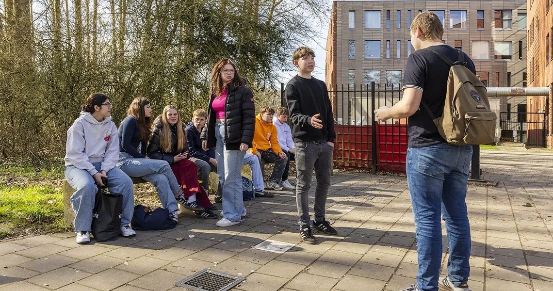 Studenten zitten en staan buiten bij een schoolgebouw terwijl een medestudent het woord voert