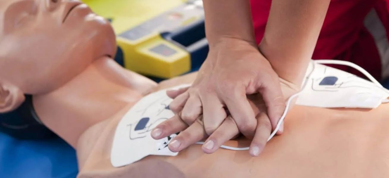 Hands performing chest compressions on a CPR training manikin with AED pads attached
