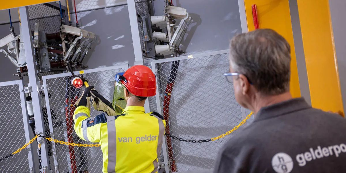 An electrical technician in protective gear working on a switchgear panel while an assessor observes