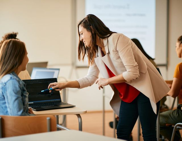 Lehrkraft, die einer Person am Laptop in einem Klassenraum hilft