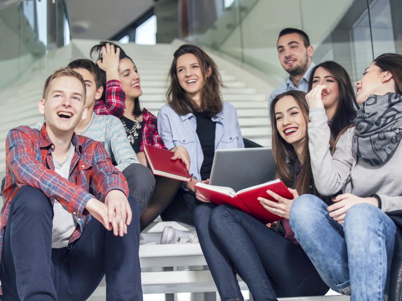 Eine Gruppe Studierender sitzt lachend mit Büchern und einem Laptop auf einer Treppe