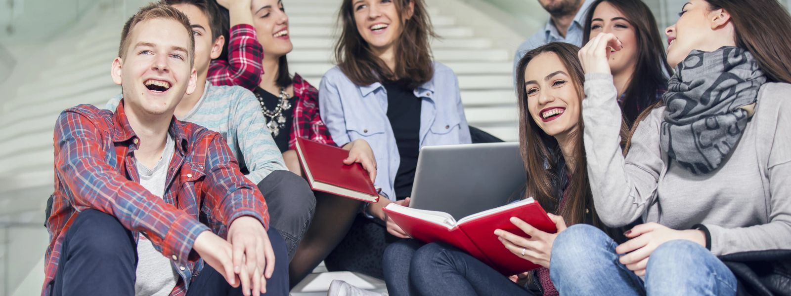A group of students sitting and laughing on a staircase with books and a laptop